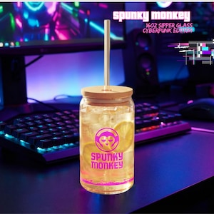 May include: A clear glass sipper with a bamboo lid and a metal straw. The glass is filled with a yellow liquid and lemon slices. The glass has a pink logo that says "Spunky Monkey". The text "16oz Sipper Glass Cyberpunk Edition" is also visible.