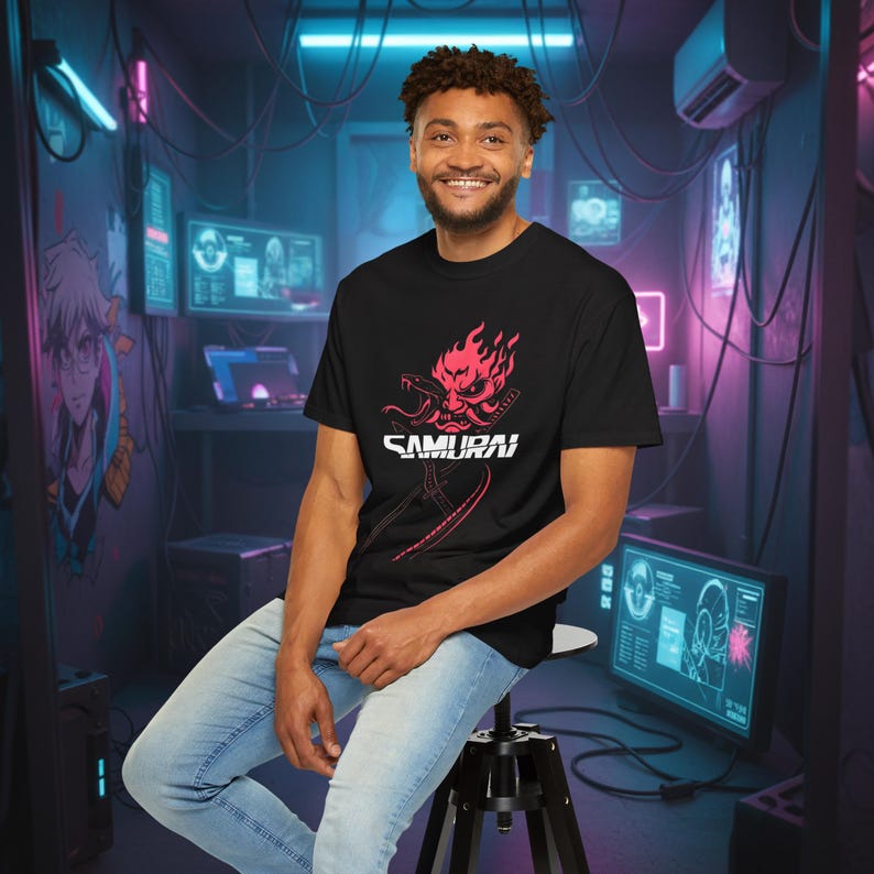 May include: Black t-shirt featuring a red and white graphic of a samurai mask with the word "SAMURAI" below. The design includes a sword and flames. The person is wearing light blue jeans and sitting on a black stool.