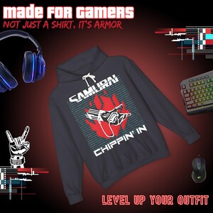 Op de afbeelding: Een donkergrijze hoodie met de tekst "SAMURAI CHIPPIN' IN" en een afbeelding van een hand en vlammen. De afbeelding bevat ook een toetsenbord, muis en koptelefoon, met de tekst "MADE FOR GAMERS" en "LEVEL UP YOUR OUTFIT".