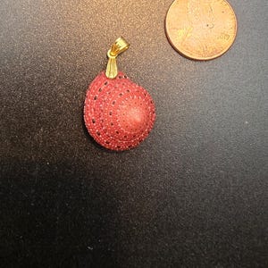May include: A pendant featuring a red, textured, spherical design with black speckles, suspended from a gold-colored bail. A penny is next to the pendant for size comparison. Jewelry.