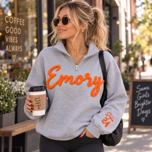 May include: A light gray quarter-zip sweatshirt featuring the word Emory in orange textured chenille lettering across the chest and the text Sofia 24 embroidered in orange on the left sleeve cuff, paired with a black shoulder bag.