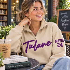 May include: A beige quarter-zip sweatshirt featuring the word Tulane in purple script embroidery across the chest and the name OLIVIA with the number 24 in purple block lettering on the left sleeve.