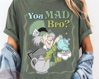 May include: Moss green t-shirt featuring a Mad Hatter graphic with the text "You MAD Bro?". The illustration depicts the Mad Hatter pouring tea. The shirt has rolled-up sleeves and a relaxed fit.