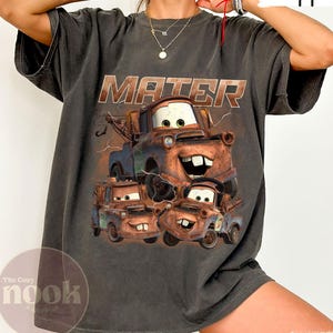 May include: Dark grey t-shirt with a graphic of Mater from the film Cars. The word "MATER" is printed above the character. The shirt has a relaxed fit and short sleeves. The Cozy Nook logo is visible.
