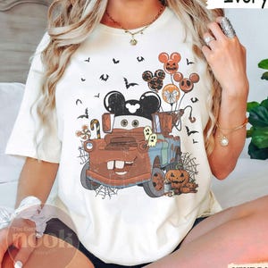 May include: Ivory-colored t-shirt featuring a Halloween-themed graphic. The design includes a cartoon truck with Mickey Mouse ears, balloons, pumpkins, ghosts, and bats. The text "Ivory" is printed on the top right.