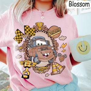 May include: Pink t-shirt featuring a cartoon image of Mater from the movie Cars, with the text "Rusteze" and "Cars". The design includes racing flags, lightning bolts, and a trophy. The shirt is being held by a person.