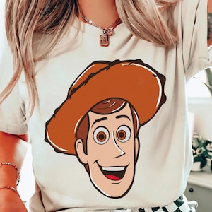 May include: A cream-colored t-shirt featuring a cartoon illustration of a smiling cowboy character with a brown hat and brown hair. The character has large brown eyes and a red mouth. The design is centered on the shirt.