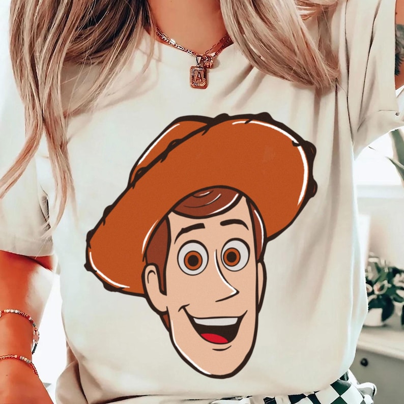 May include: A cream-colored t-shirt featuring a cartoon illustration of a smiling cowboy character with a brown hat and brown hair. The character has large brown eyes and a red mouth. The design is centered on the shirt.