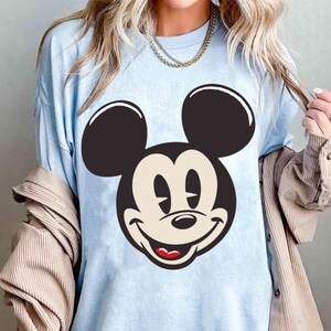 May include: Light blue t-shirt featuring a large graphic of Mickey Mouse's face. The design includes black ears, a beige face, and a red mouth. The shirt has a relaxed fit and short sleeves.