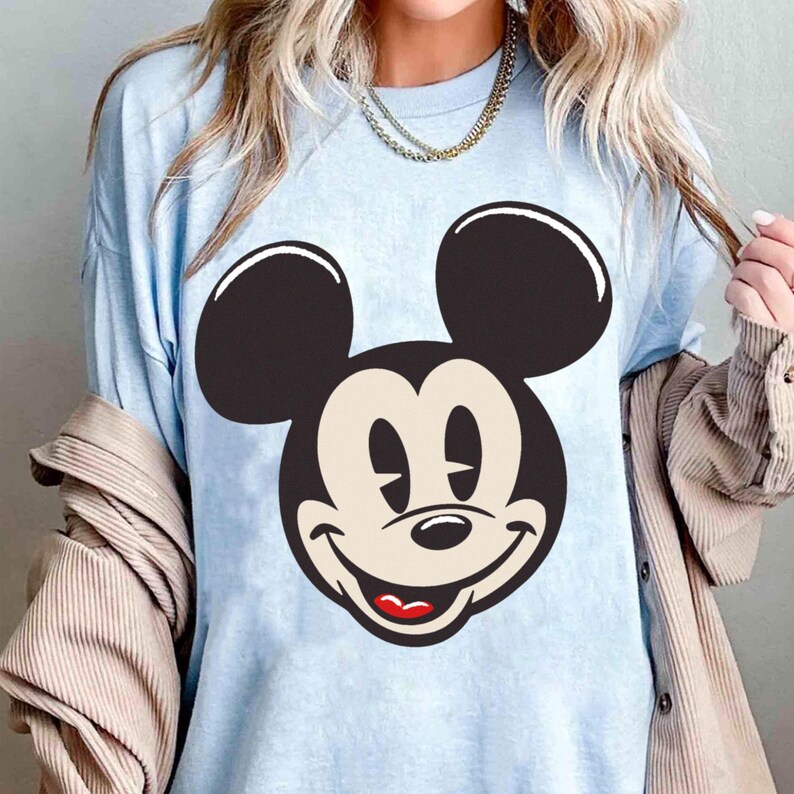 May include: Light blue t-shirt featuring a large graphic of Mickey Mouse's face. The design includes black ears, a beige face, and a red mouth. The shirt has a relaxed fit and short sleeves.