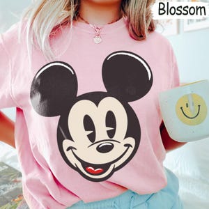 May include: A light pink sweatshirt featuring a large graphic of Mickey Mouse's face in black, beige, and red. The word "Blossom" is in the top right corner. A white mug with a yellow smiley face is held in the image.