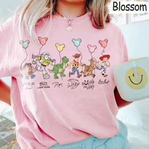 May include: Pale pink t-shirt with a cartoon illustration of Toy Story characters. The design includes Potato Head, Buzz Lightyear, Rex, Woody, Bullseye, and Bo Peep, each with a balloon. The word "Blossom" is visible.