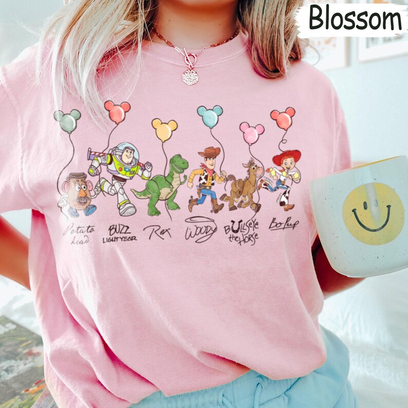 May include: Pale pink t-shirt with a cartoon illustration of Toy Story characters. The design includes Potato Head, Buzz Lightyear, Rex, Woody, Bullseye, and Bo Peep, each with a balloon. The word "Blossom" is visible.