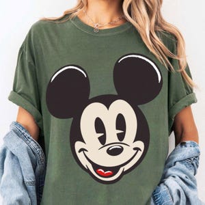 May include: Olive green t-shirt featuring a large graphic of Mickey Mouse's face. The cartoon character's face is black, white, and red. The shirt has short sleeves and a relaxed fit.