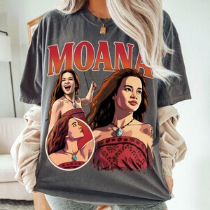 以下が含まれることがあります： チャコールグレーの半袖Tシャツで、キャラクター「モアナ」の大きなグラフィックプリントが施されています。デザインには太い赤文字で「MOANA」と書かれており、青いペンダントネックレスを身に着けたキャラクターの3つの異なるイラストが描かれています。