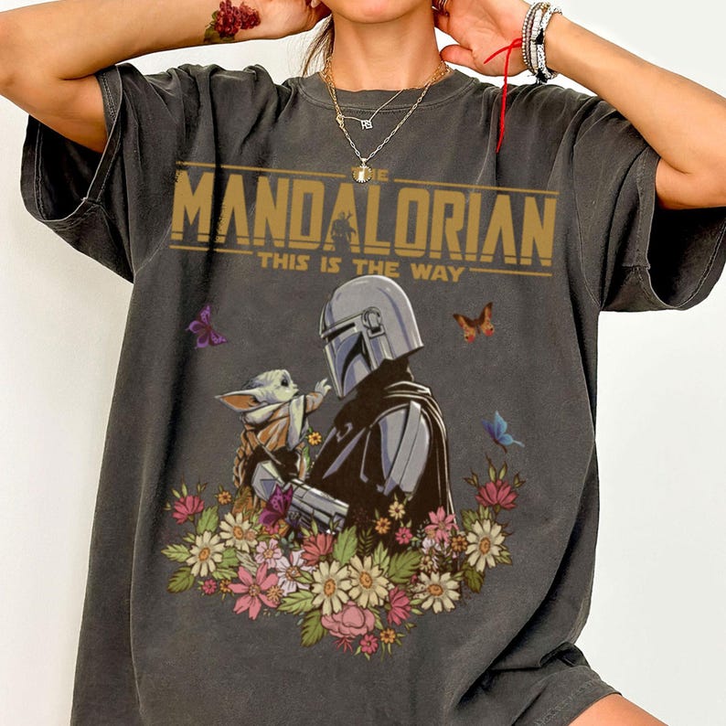 May include: Dark gray t-shirt featuring a graphic of The Mandalorian and Grogu surrounded by flowers and butterflies. The text "THE MANDALORIAN" and "THIS IS THE WAY" are printed across the top in gold.