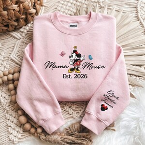 K&ouml;nnte beinhalten: Hellrosa Sweatshirt mit einem Minnie Mouse-Design und dem Text "Mama Mouse Est. 2026". Der &Auml;rmel zeigt gestickte Namen. Das Design umfasst Schmetterlinge und eine Minnie Mouse-Figur.
