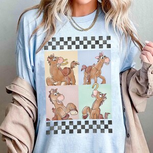 May include: Light blue t-shirt featuring a four-panel design of a cartoon horse with a saddle and a horseshoe. The horse is brown with a pink nose and a smiling expression. The top and bottom of the design have a black and white checkered pattern.