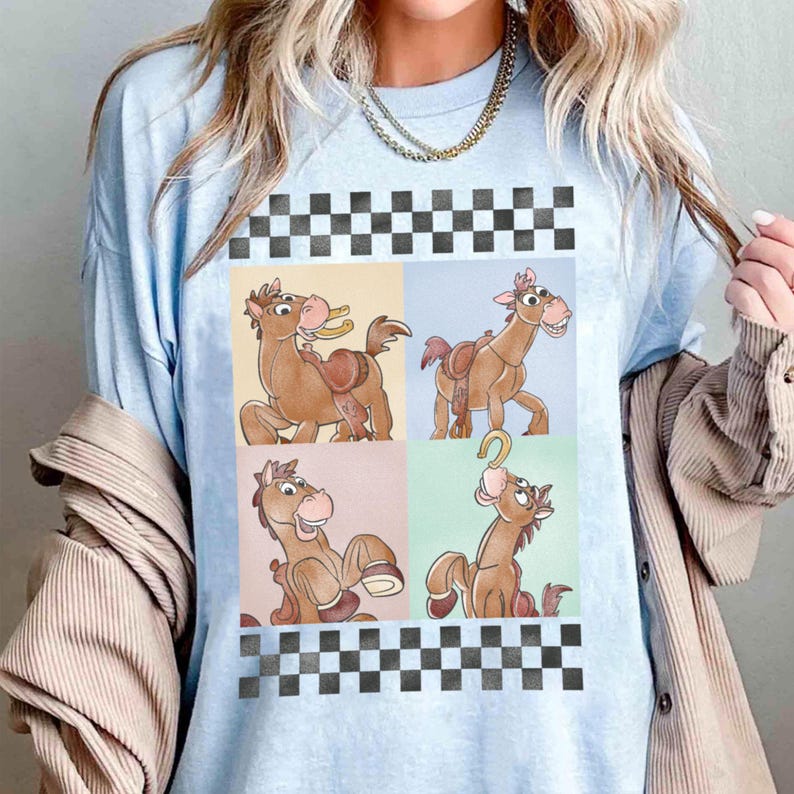 May include: Light blue t-shirt featuring a four-panel design of a cartoon horse with a saddle and a horseshoe. The horse is brown with a pink nose and a smiling expression. The top and bottom of the design have a black and white checkered pattern.