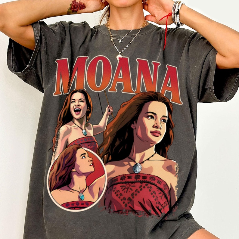 以下が含まれることがあります： 大きな赤い文字でMOANAと書かれたチャコールグレーの半袖Tシャツ。グラフィックには、赤い柄のトップスと青いペンダントネックレスを身に着けたキャラクター、モアナのイラストが3つ、暗い背景に描かれています。