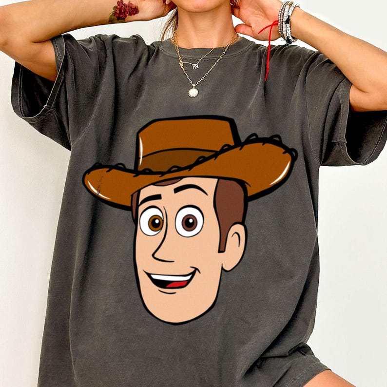 May include: Dark gray t-shirt featuring a cartoon illustration of a cowboy's face with a brown hat. The cowboy has brown hair, brown eyes, and a smiling expression. The shirt is a casual style.