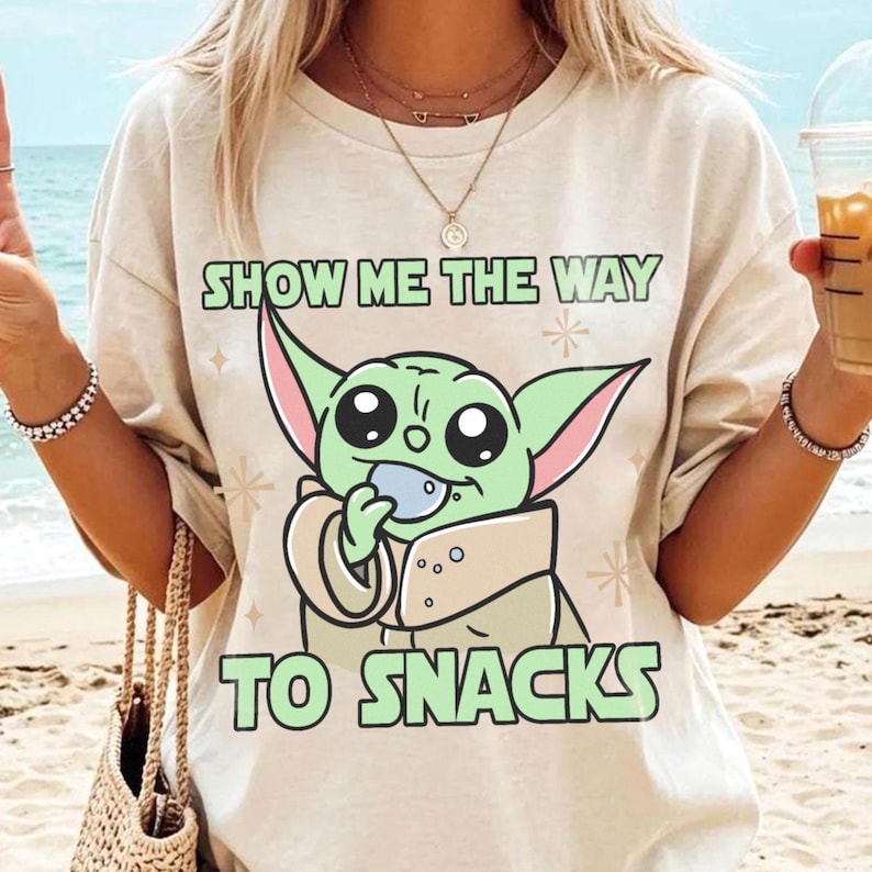 Peut inclure: T-shirt beige avec un personnage de dessin anim&eacute; vert tenant un objet bleu. Le texte "SHOW ME THE WAY TO SNACKS" est imprim&eacute; au-dessus et en dessous du personnage en vert. Le t-shirt est port&eacute; par une personne sur une plage.