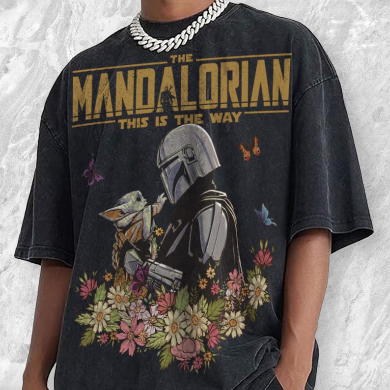 May include: Black t-shirt featuring a graphic of The Mandalorian and Grogu surrounded by flowers and butterflies. The text "THE MANDALORIAN THIS IS THE WAY" is printed across the top in gold lettering. The shirt has a vintage, distressed look.