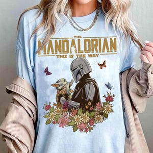 May include: Light blue t-shirt featuring a graphic of The Mandalorian and Grogu surrounded by flowers and butterflies. The text "THE MANDALORIAN" and "THIS IS THE WAY" are printed above the graphic.
