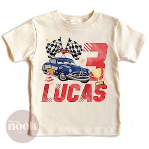 May include: Cream-colored t-shirt featuring a blue cartoon car with the text "Fabulous Hudson Hornet" and the name "Lucas" in red. Includes a number "3", checkered flags, and a gold trophy.
