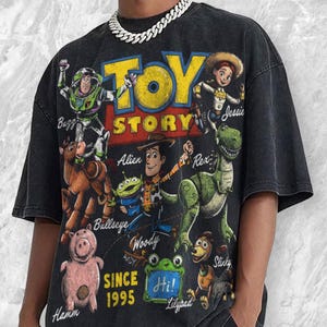 Pode incluir: Uma t-shirt preta de manga curta com uma grande impress&atilde;o gr&aacute;fica de personagens de Toy Story, incluindo Buzz, Jessie, Woody, Rex, Bullseye, Hamm e um Alien. O texto diz Toy Story, Since 1995 e Hi! Lilypad. Acabamento de lavagem vintage.