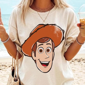 May include: A beige t-shirt featuring a cartoon illustration of a smiling cowboy character with a brown hat and hair, and brown eyes. The shirt is a casual style, suitable for everyday wear.