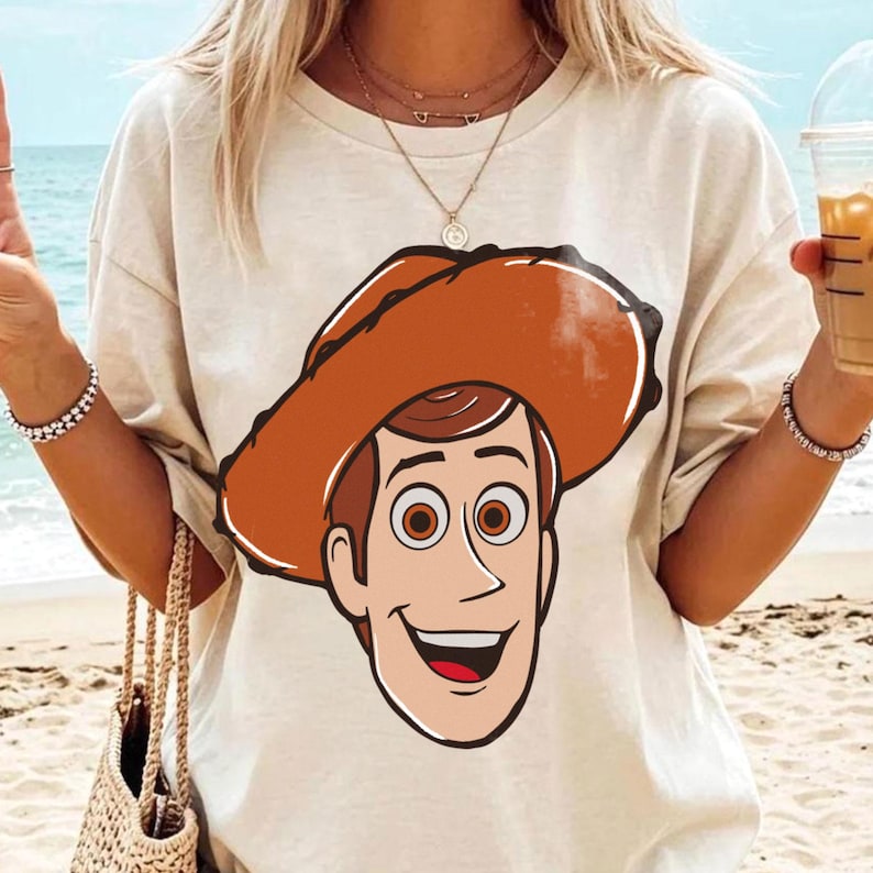 May include: A beige t-shirt featuring a cartoon illustration of a smiling cowboy character with a brown hat and hair, and brown eyes. The shirt is a casual style, suitable for everyday wear.