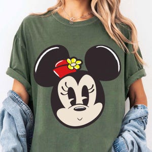 Peut inclure: T-shirt vert olive avec un grand dessin du visage de Minnie Mouse. Le visage est de couleur cr&egrave;me avec des oreilles, des cils et un petit sourire noirs. Elle porte un n&oelig;ud rouge avec une fleur jaune. Le t-shirt a des manches courtes.