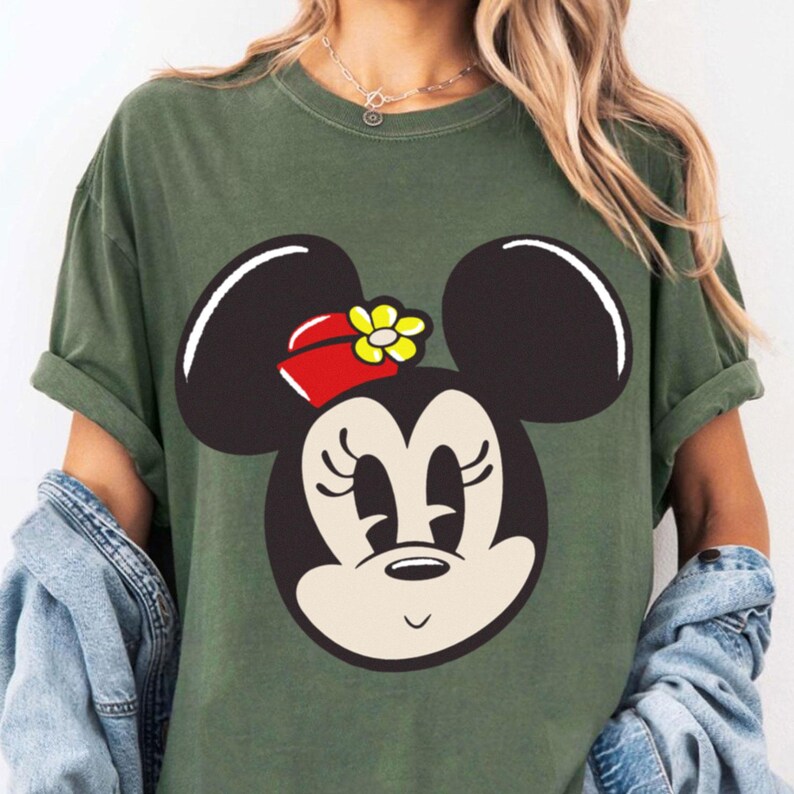 Peut inclure: T-shirt vert olive avec un grand dessin du visage de Minnie Mouse. Le visage est de couleur cr&egrave;me avec des oreilles, des cils et un petit sourire noirs. Elle porte un n&oelig;ud rouge avec une fleur jaune. Le t-shirt a des manches courtes.