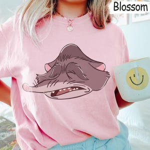 Peut inclure: Un sweat-shirt rose clair avec une illustration de dessin anim&eacute; d'une cr&eacute;ature grise endormie, les yeux ferm&eacute;s et la bouche l&eacute;g&egrave;rement ouverte. Le mot "Blossom" est imprim&eacute; en haut &agrave; droite. Une tasse blanche avec un smiley jaune est tenue.