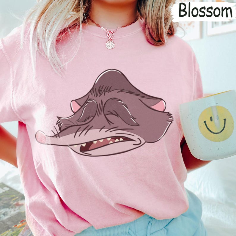 Peut inclure: Un sweat-shirt rose clair avec une illustration de dessin anim&eacute; d'une cr&eacute;ature grise endormie, les yeux ferm&eacute;s et la bouche l&eacute;g&egrave;rement ouverte. Le mot "Blossom" est imprim&eacute; en haut &agrave; droite. Une tasse blanche avec un smiley jaune est tenue.