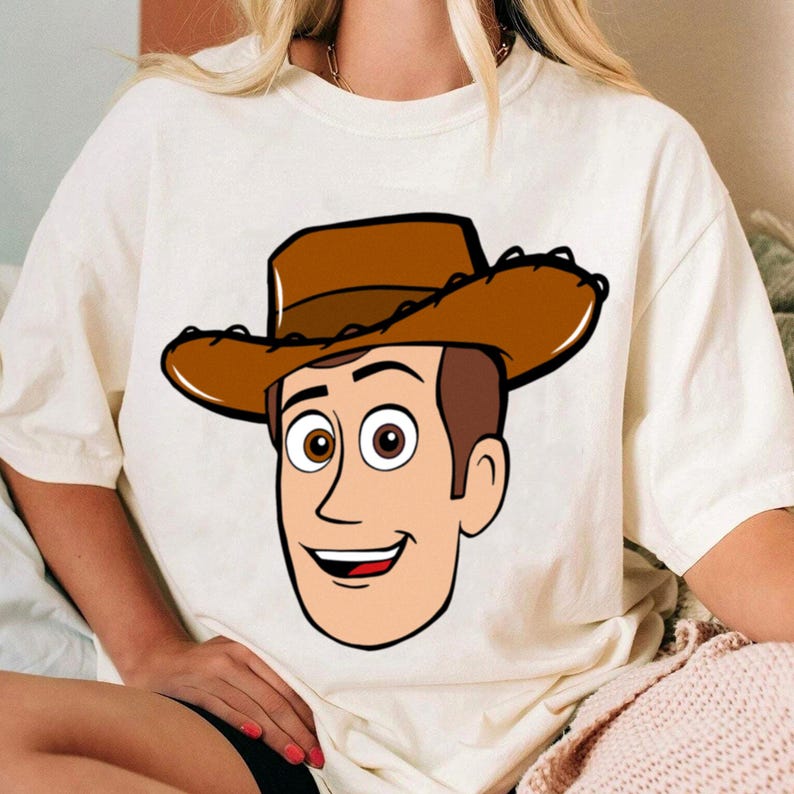 May include: Cream-colored t-shirt featuring a cartoon illustration of a smiling cowboy character wearing a brown hat. The graphic is a detailed, outlined design with brown hair and eyes, and a red mouth.