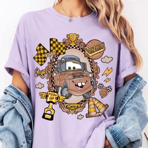 May include: A light purple t-shirt featuring a cartoon image of Mater from the movie Cars, framed in a decorative border. The design includes racing flags, lightning bolts, and the words "Rusteze" and "Cars".