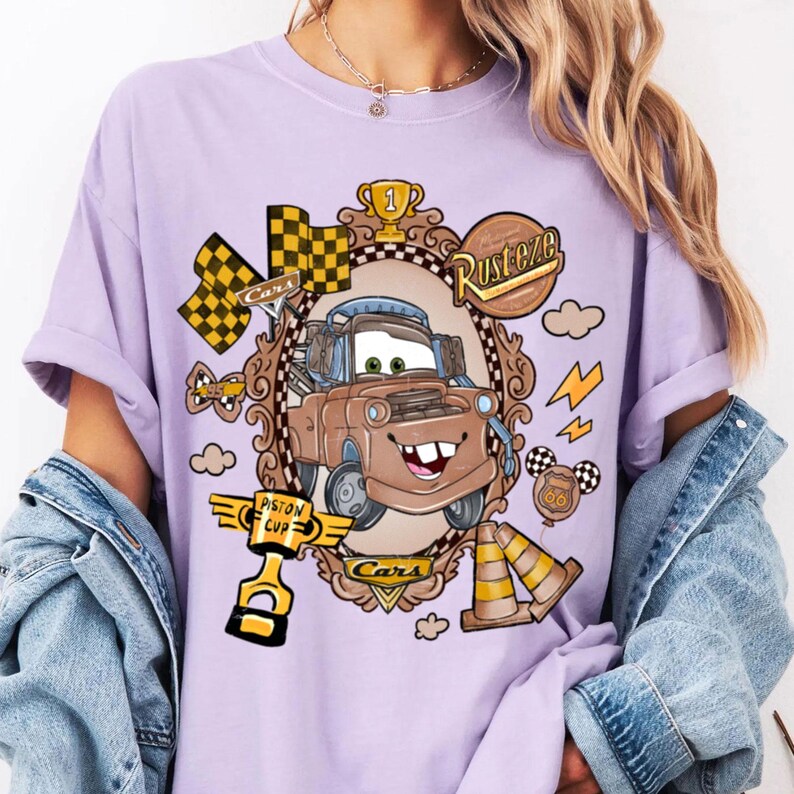 May include: A light purple t-shirt featuring a cartoon image of Mater from the movie Cars, framed in a decorative border. The design includes racing flags, lightning bolts, and the words "Rusteze" and "Cars".