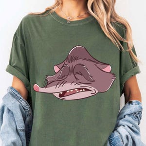 Peut inclure: T-shirt vert olive avec une illustration de dessin anim&eacute; d'une cr&eacute;ature grincheuse aux yeux ferm&eacute;s, au nez rose et &agrave; la bouche ouverte. Le t-shirt a des manches courtes et une coupe d&eacute;contract&eacute;e.