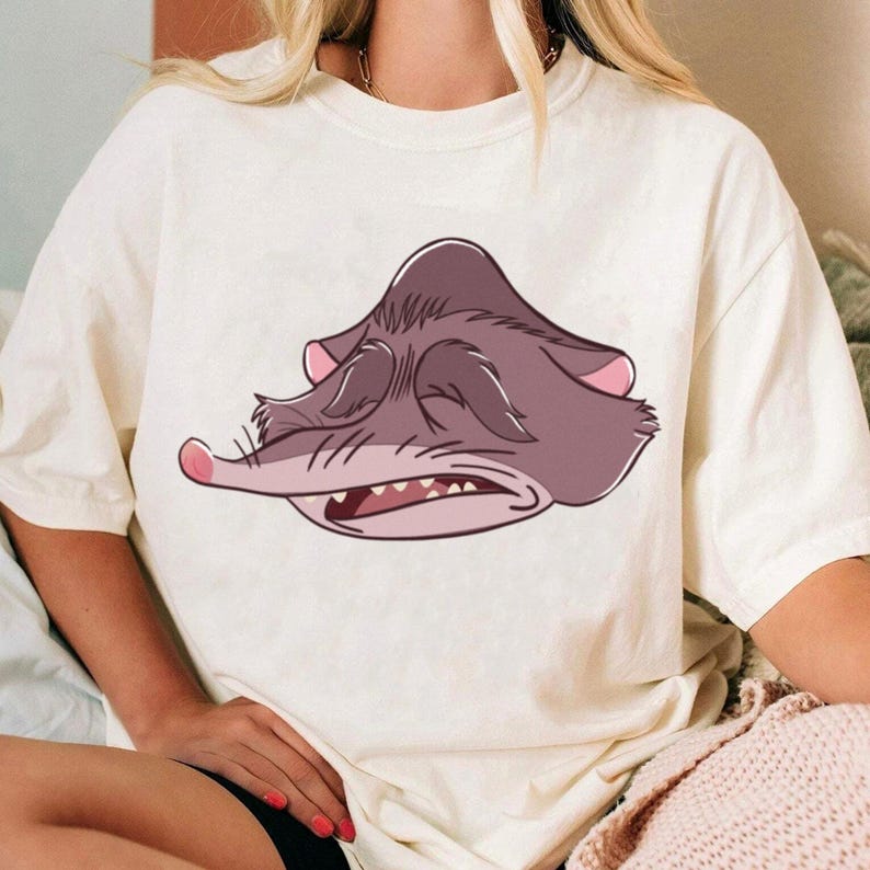 Peut inclure: T-shirt cr&egrave;me avec un visage de possum de dessin anim&eacute; aux yeux ferm&eacute;s et &agrave; la bouche ouverte, montrant les dents. Le possum est principalement dans les tons de violet et de rose. Le t-shirt est de style d&eacute;contract&eacute; et confortable.