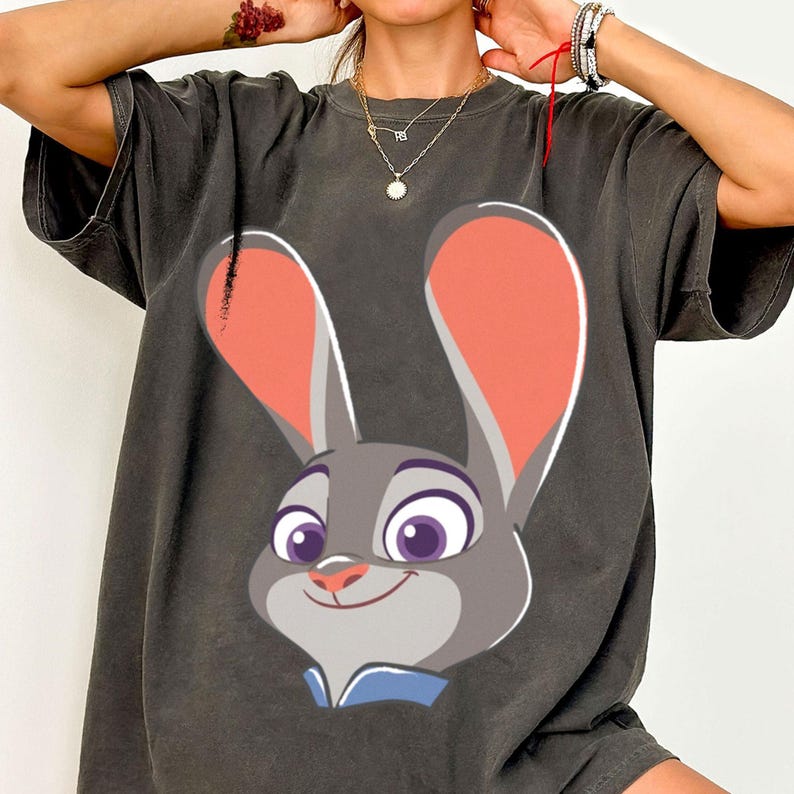 Puede incluir: Camiseta gris oscuro con una imagen de dibujos animados de Judy Hopps de Zootr&oacute;polis. El gr&aacute;fico muestra la cara de Judy con grandes orejas grises, interiores naranjas y ojos morados. La camiseta tiene cuello redondo y mangas cortas.