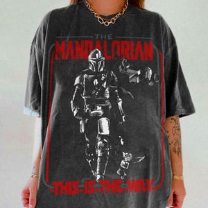 May include: Dark gray t-shirt featuring a graphic of The Mandalorian, a character from the Star Wars series. The design includes the text "THE MANDALORIAN" in red, and "THIS IS THE WAY" in red. The image also shows a small figure of Grogu.