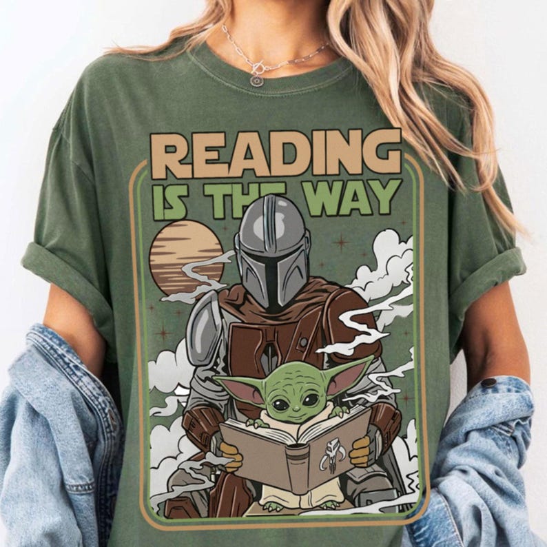 Puede incluir: Camiseta verde con el texto "READING IS THE WAY" en un estilo retro. El dise&ntilde;o gr&aacute;fico presenta un personaje mandaloriano y Grogu leyendo un libro, con un planeta y nubes de fondo.