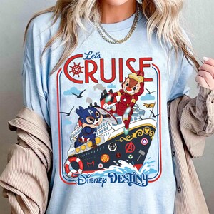 May include: Light blue t-shirt with the words "Let's Cruise" in red. The shirt features a graphic of a cruise ship with cartoon characters dressed as superheroes. The ship has the words "Disney Destiny" below it.