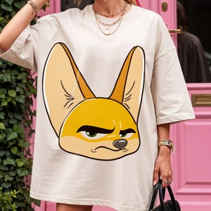 May include: Oversized, cream-colored t-shirt featuring a cartoon illustration of a fennec fox. The fox has large, pointed ears, a yellow-orange face, and a grumpy expression. The shirt is a casual, comfortable style.