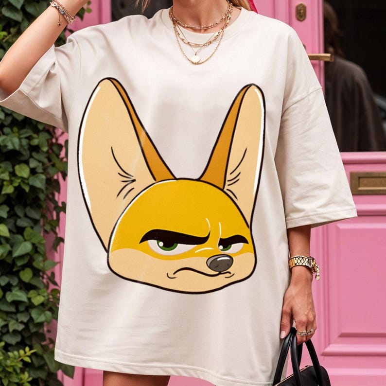 May include: Oversized, cream-colored t-shirt featuring a cartoon illustration of a fennec fox. The fox has large, pointed ears, a yellow-orange face, and a grumpy expression. The shirt is a casual, comfortable style.