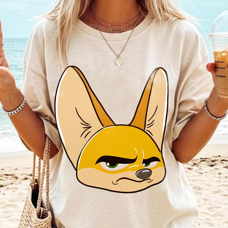 May include: A beige t-shirt featuring a cartoon illustration of a fennec fox with large ears and a grumpy expression. The fox is primarily yellow and tan with green eyes. The shirt is worn by a person on a beach.