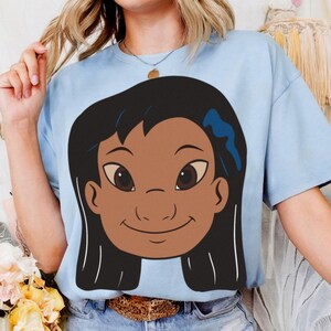 Puede incluir: Camiseta azul claro con una ilustraci&oacute;n de dibujos animados de una ni&ntilde;a sonriente con cabello oscuro y ojos marrones. La ni&ntilde;a tiene un lazo azul en el pelo. La camiseta est&aacute; hecha de un material suave y tiene mangas cortas.