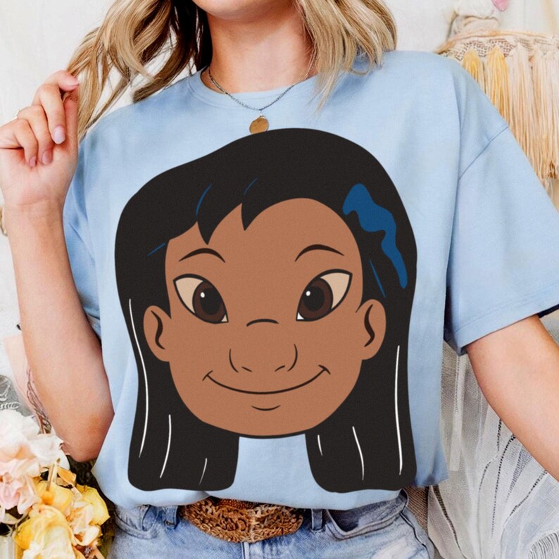 Puede incluir: Camiseta azul claro con una ilustraci&oacute;n de dibujos animados de una ni&ntilde;a sonriente con cabello oscuro y ojos marrones. La ni&ntilde;a tiene un lazo azul en el pelo. La camiseta est&aacute; hecha de un material suave y tiene mangas cortas.